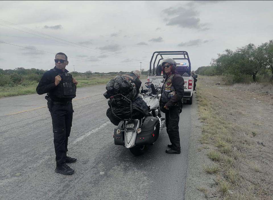 Two law enforcement officers stand beside a motorcycle stopped on a rural highway, with a police cruiser and flashing lights in the background.